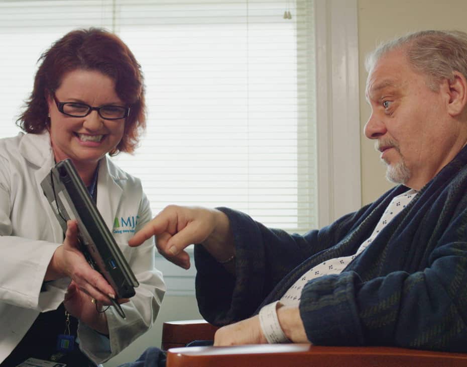 Una cuidadora de MJHS sostiene sonriendo una tableta para un hombre mayor