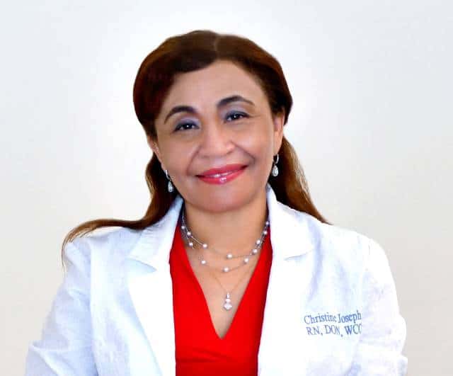 A female-presenting person with long brown hair pulled back from her face, wearing a white lab coat over a red top, smiles at the camera