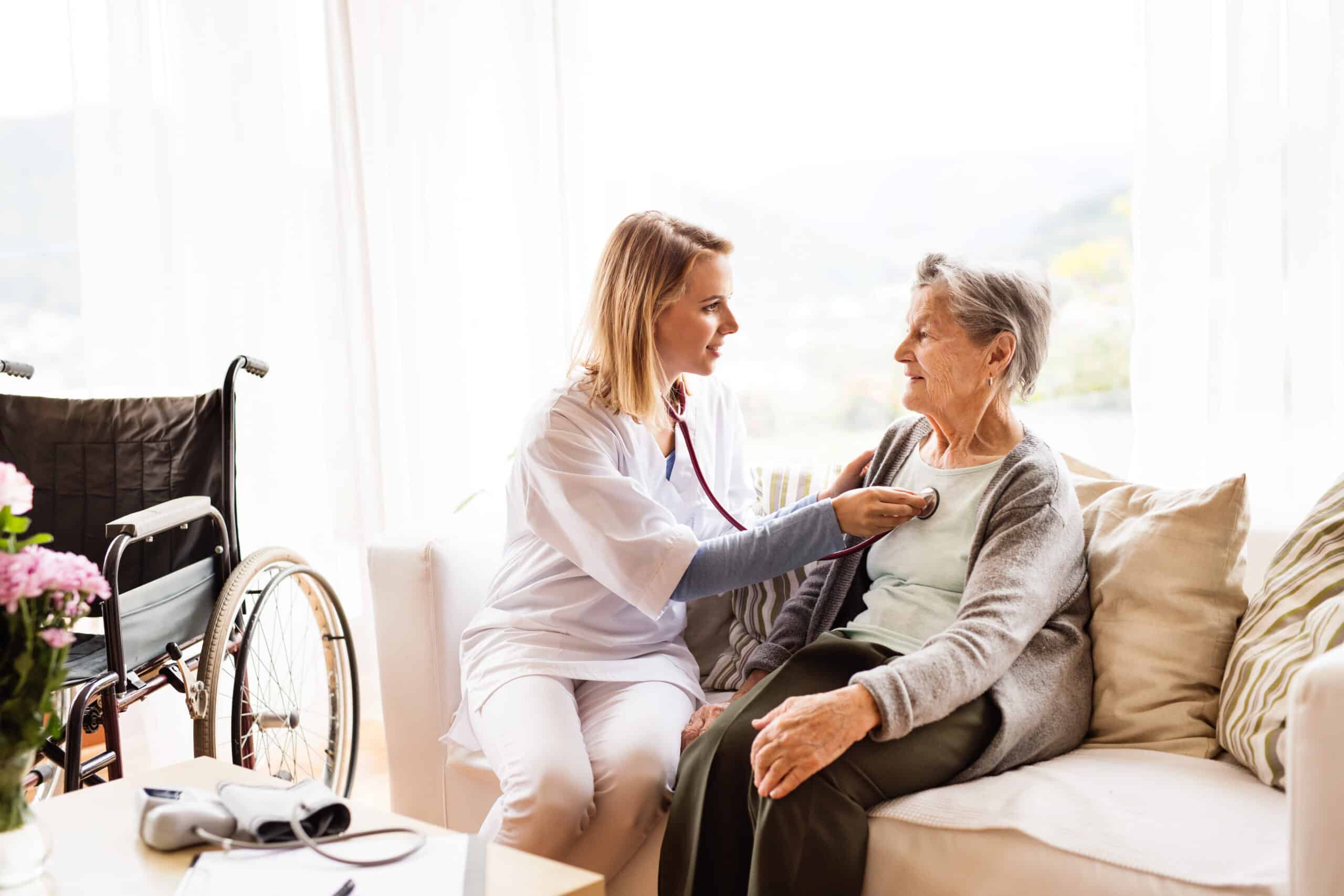 A home health nurse sits with a patient on a couch. She has a stethoscope and is listening to the patient's heart.